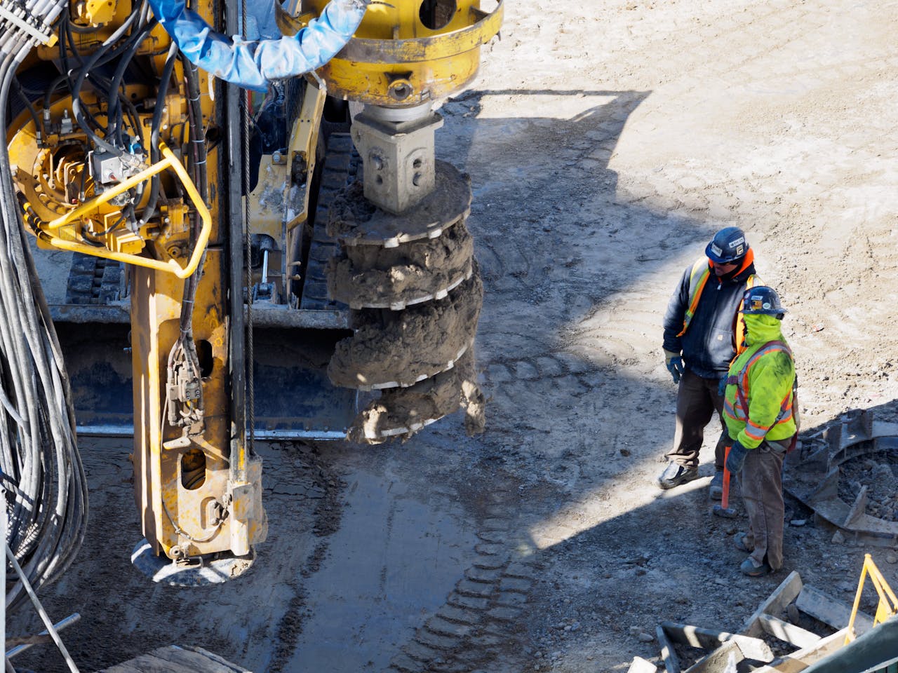 Two workers in protective gear at a construction site with heavy drilling machinery.